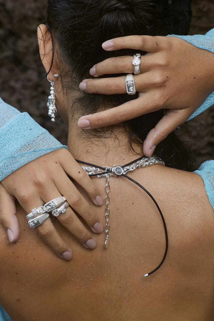 Sculptural and serene, the Maia Ring in silver brings together ancient influence and modern clarity. Its ribbed circular band is crafted in a cool silver-tone finish, framing two rectangular cubic zirconia stones in molten-textured bezel settings—subtle sparkles that evoke the shimmer of starlight on water.