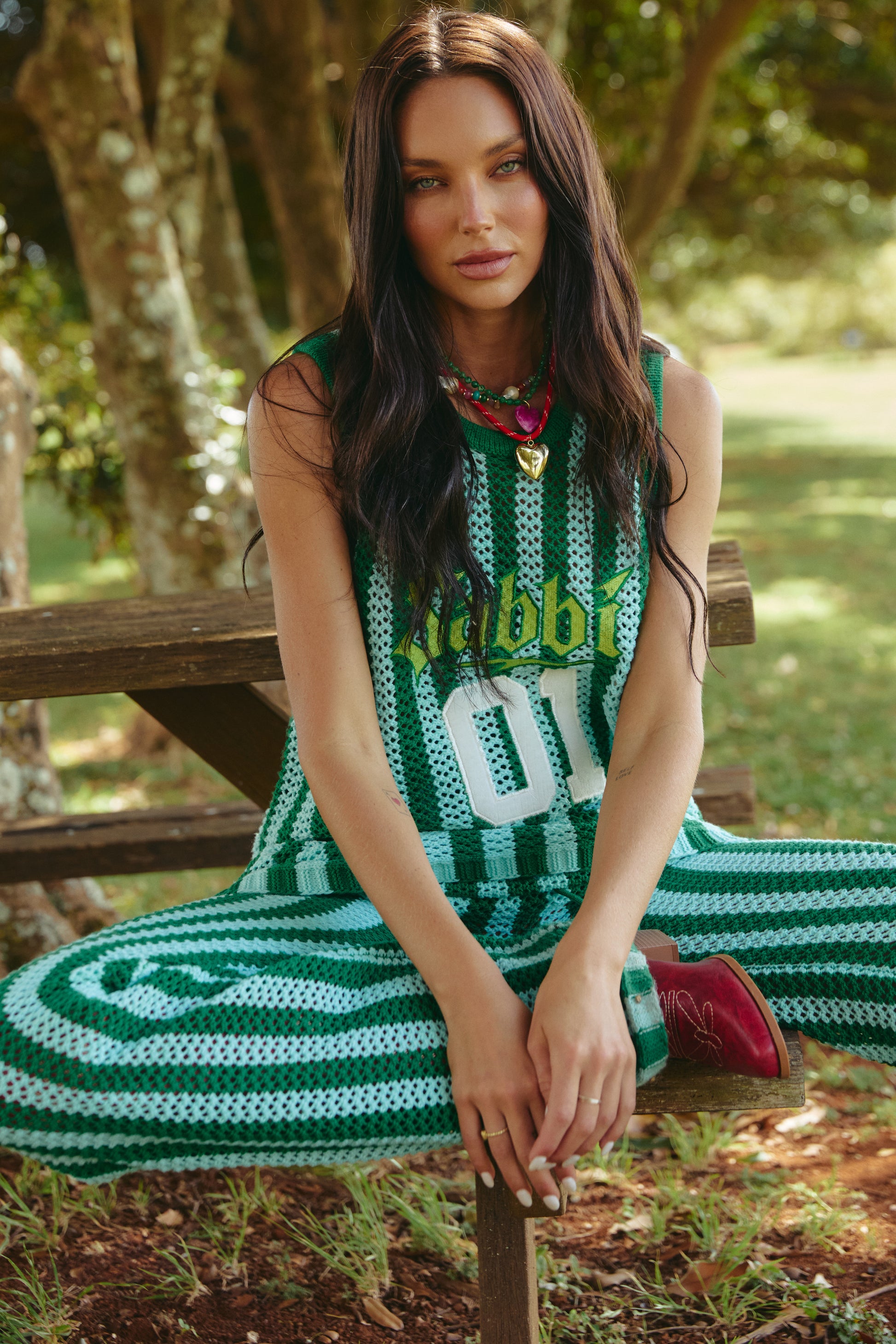 A woman with long dark hair sits on a wooden bench outdoors, wearing a green and white striped outfit and red shoes. The scene is calm and natural.