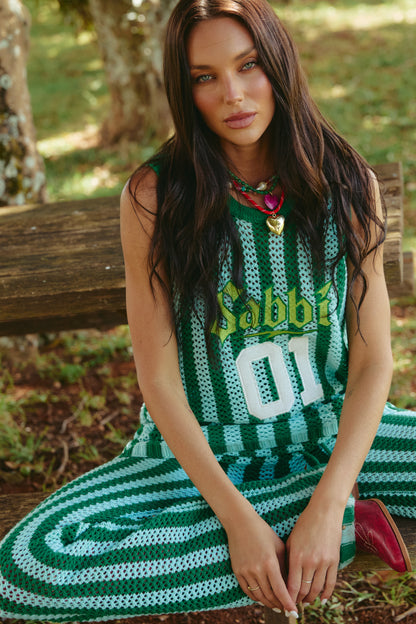 Woman with long dark hair sits cross-legged on a wooden bench outdoors. She wears a green and white striped outfit with "Sabbia" and "01" on the top.