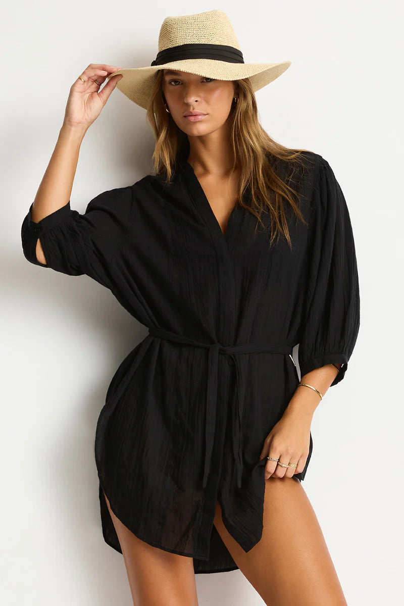 A woman in a black belted dress and broad-brimmed straw hat poses confidently against a light background. She exudes a relaxed, chic summer vibe.