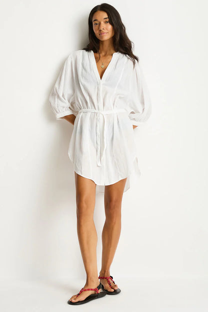A woman in a white, flowy shirt dress with a tie belt stands against a plain background. She wears red sandals. The mood appears relaxed and casual.