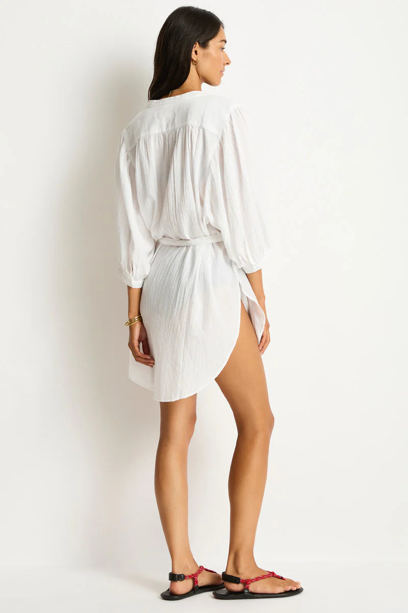 A woman in a flowy, white, long-sleeved dress stands sideways, highlighting the dress's texture. She wears red sandals on a plain background.